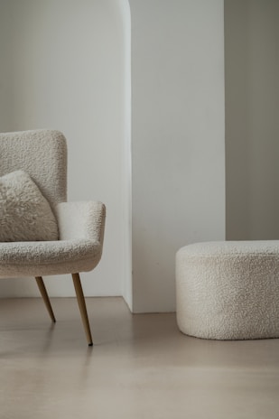 A modern, minimalist room featuring a cozy armchair with a fluffy cushion and a matching round ottoman. Both furniture pieces have a soft, textured fabric in a light cream color. They are set against a light-colored wall with a unique architectural design.