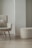 A modern, minimalist room featuring a cozy armchair with a fluffy cushion and a matching round ottoman. Both furniture pieces have a soft, textured fabric in a light cream color. They are set against a light-colored wall with a unique architectural design.