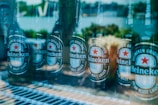 Several Heineken beer bottles are arranged next to each other on a shelf, with a reflection of an outdoor setting visible through the glass. The labels are clearly visible, displaying the brand and logo.