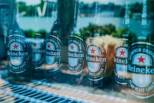 Several Heineken beer bottles are arranged next to each other on a shelf, with a reflection of an outdoor setting visible through the glass. The labels are clearly visible, displaying the brand and logo.