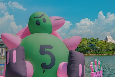 A large inflatable green and pink object resembling a turtle with a number five printed on it is situated near a body of water. In the background, there are buildings and greenery under a sunny sky with fluffy white clouds.