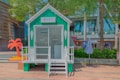 A small green and white kiosk is situated outdoors on a paved area. The kiosk has a triangular roof, a sliding glass door, and a small staircase leading up to it. Nearby, there is a decorative orange palm tree and a blue and white striped umbrella. Trees with green foliage are seen in the background, along with other modern buildings and structures.