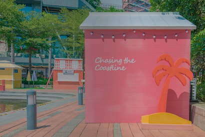 A pink wooden structure with a gray roof is adorned with string lights and features the words 'Chasing the Coastline' in white script. Next to the text is a stylized orange palm tree with a yellow base. The scene includes part of a walkway, a small water feature, and other colorful structures. There are trees and a modern building in the background.