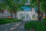 A modern architectural structure surrounded by lush trees and greenery, with a building featuring large glass panels in the background. The foreground includes a paved path lined with plants and trees, creating a harmonious blend of nature and urban design.