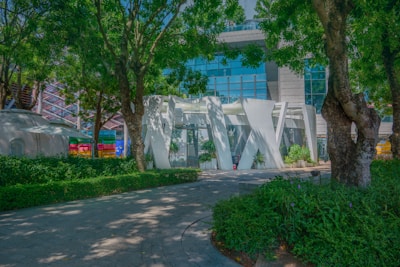 A modern architectural structure surrounded by lush trees and greenery, with a building featuring large glass panels in the background. The foreground includes a paved path lined with plants and trees, creating a harmonious blend of nature and urban design.
