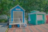 A small vibrant blue cabin with a porch and staircase sits next to a teal booth. The cabin has a white trim and a sign above the door. An easel is placed at the entrance. Two closed umbrellas with checkered patterns stand nearby. The area is surrounded by lush green trees and a curved, paved path.