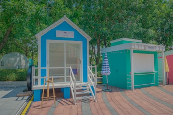A small vibrant blue cabin with a porch and staircase sits next to a teal booth. The cabin has a white trim and a sign above the door. An easel is placed at the entrance. Two closed umbrellas with checkered patterns stand nearby. The area is surrounded by lush green trees and a curved, paved path.