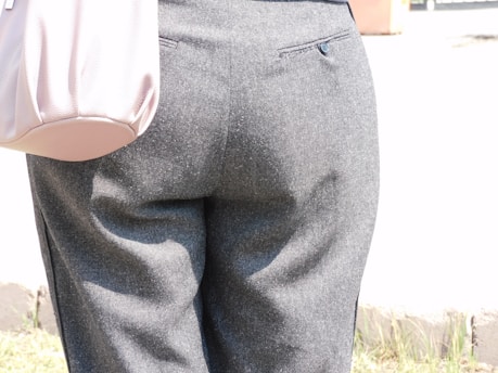 A close-up view of a person wearing gray pants with a pocket detail is visible at the back. The material appears to be wool or a similar fabric, and the pants are tailored with a buttoned pocket. A light pink bag is partially visible on the left side.