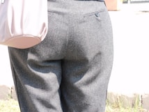 A close-up view of a person wearing gray pants with a pocket detail is visible at the back. The material appears to be wool or a similar fabric, and the pants are tailored with a buttoned pocket. A light pink bag is partially visible on the left side.