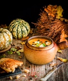 A rustic scene featuring a ceramic bowl filled with pumpkin soup, garnished with seeds and herbs. Surrounding the bowl are autumn elements like small pumpkins, dry leaves, and a bundle of dry twigs. A piece of crusty bread with a bite taken out lies near a knife on a wooden surface.