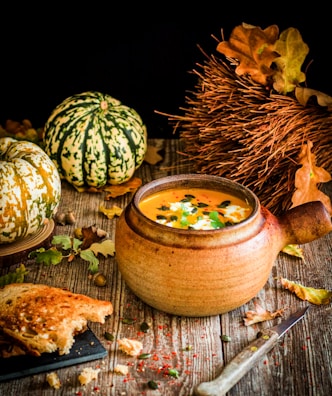 A rustic scene featuring a ceramic bowl filled with pumpkin soup, garnished with seeds and herbs. Surrounding the bowl are autumn elements like small pumpkins, dry leaves, and a bundle of dry twigs. A piece of crusty bread with a bite taken out lies near a knife on a wooden surface.