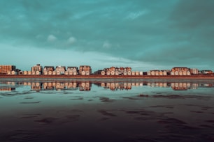 A row of charming seaside houses is reflected in the calm water of a beach at low tide. The sky is overcast with a mix of gray and teal clouds, enhancing the tranquil mood. The houses are mostly white with dark roofs and are lined up side by side, creating a picturesque coastal scene.