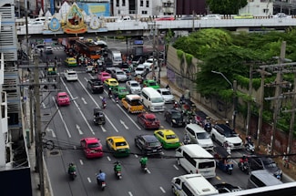 A busy urban street with heavy traffic, including a variety of vehicles such as cars, taxis in pink and green, buses, and numerous motorbikes. Elevated walkways and lush greenery are visible, along with power lines and buildings. The atmosphere is bustling with people and vehicles moving in different directions.