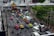 A busy urban street with heavy traffic, including a variety of vehicles such as cars, taxis in pink and green, buses, and numerous motorbikes. Elevated walkways and lush greenery are visible, along with power lines and buildings. The atmosphere is bustling with people and vehicles moving in different directions.