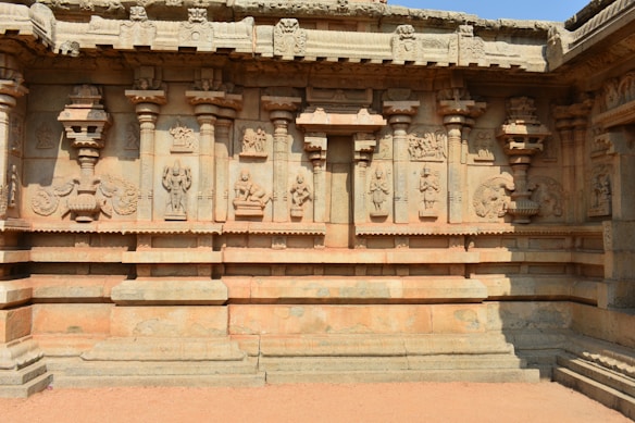 A detailed stone wall featuring intricate carvings and sculptures depicting figures and ornamental designs. The wall includes several columns and pilasters with detailed motifs and images, reflecting ancient architectural artistry.