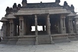 An ancient stone structure featuring intricate carvings and pillars, suggesting a historic temple or monument. The architecture includes detailed engravings and multiple columns, evoking a sense of antiquity and grandeur.