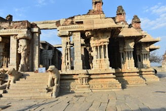 Ancient stone temple structure featuring intricately carved pillars and figures. The architecture showcases detailed sculptural art, with steps leading up to the entrance flanked by animal sculptures. The temple is partially weathered, suggesting historical significance and age.