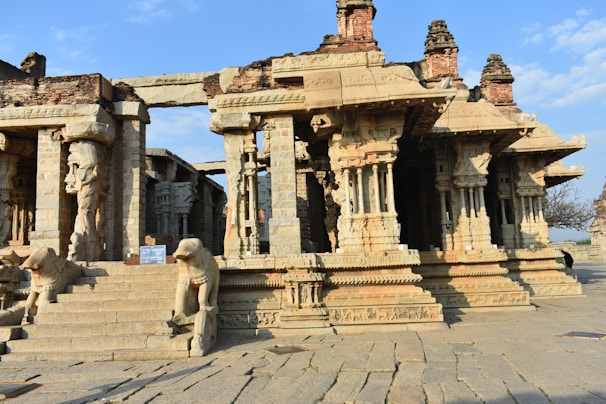 Ancient stone temple structure featuring intricately carved pillars and figures. The architecture showcases detailed sculptural art, with steps leading up to the entrance flanked by animal sculptures. The temple is partially weathered, suggesting historical significance and age.