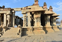 Ancient stone temple structure featuring intricately carved pillars and figures. The architecture showcases detailed sculptural art, with steps leading up to the entrance flanked by animal sculptures. The temple is partially weathered, suggesting historical significance and age.