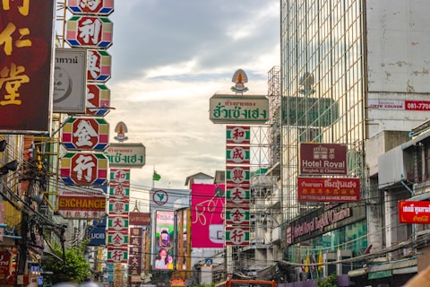 A bustling urban street scene features colorful signage in multiple languages, predominantly with Asian characters, indicating a multicultural area. The environment feels dense with various wires crisscrossing above and a reflection of the cityscape visible in the glass facade of a tall building on the right.
