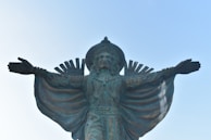 A large statue of a figure with outstretched arms, wearing ornate traditional attire with intricate detailing. The statue includes a headdress with spikes and textiles that appear draped over the arms, extending in a wide arc.
