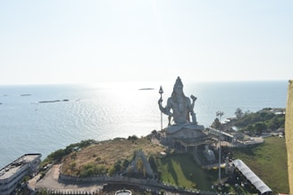 A large statue of a deity with a trident overlooks the ocean. The statue is situated on a grassy hill surrounded by a fence. Several small structures and paths are visible around the statue, indicating a temple complex. The ocean is calm, with small waves gently lapping against distant rocks under a bright sky.