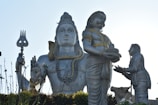 A large statue depicting a significant Hindu deity is prominently displayed. The figure is adorned with intricate jewelry and holds a trident. An elaborately coiled snake is wrapped around its neck. Two other figures are positioned in front, one offering a plate with objects on it, and a cow is present in the composition.