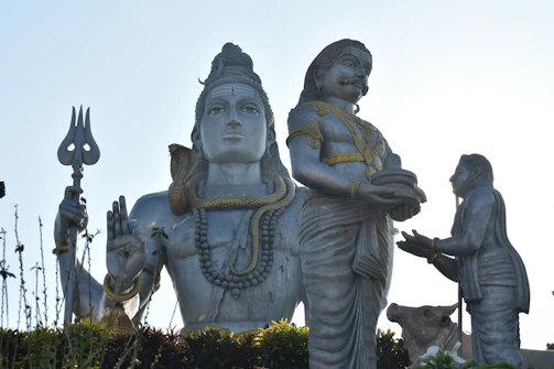 A large statue depicting a significant Hindu deity is prominently displayed. The figure is adorned with intricate jewelry and holds a trident. An elaborately coiled snake is wrapped around its neck. Two other figures are positioned in front, one offering a plate with objects on it, and a cow is present in the composition.
