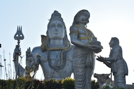 A large statue depicting a significant Hindu deity is prominently displayed. The figure is adorned with intricate jewelry and holds a trident. An elaborately coiled snake is wrapped around its neck. Two other figures are positioned in front, one offering a plate with objects on it, and a cow is present in the composition.
