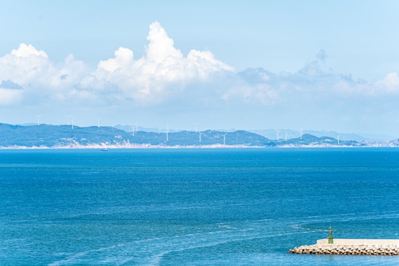 A tranquil seascape with a vast expanse of blue water in the foreground. In the distance, there is a coastline adorned with numerous wind turbines, indicating an offshore wind farm. The sky above is bright blue with large, fluffy white clouds, suggesting a clear day. On the right, a rocky pier with a small green navigational light extends into the water.