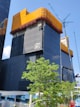 A modern high-rise building under construction with a dark glass facade. The top section is wrapped in orange mesh material, possibly for protection or safety. A tall construction crane is visible on the right side, next to the building. In the foreground, a lush green tree adds contrast to the urban setting. The sky is clear and blue.