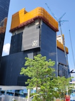 A modern high-rise building under construction with a dark glass facade. The top section is wrapped in orange mesh material, possibly for protection or safety. A tall construction crane is visible on the right side, next to the building. In the foreground, a lush green tree adds contrast to the urban setting. The sky is clear and blue.