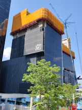 A modern high-rise building under construction with a dark glass facade. The top section is wrapped in orange mesh material, possibly for protection or safety. A tall construction crane is visible on the right side, next to the building. In the foreground, a lush green tree adds contrast to the urban setting. The sky is clear and blue.