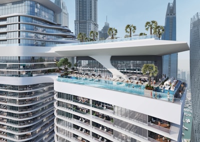 A modern skyscraper features a luxurious rooftop with an expansive infinity pool and lounging area. The building's curved glass and steel design is surrounded by tall city skyscrapers. Palm trees and sun loungers are scattered around the pool, where people are relaxing and enjoying the view.
