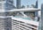 A modern skyscraper features a luxurious rooftop with an expansive infinity pool and lounging area. The building's curved glass and steel design is surrounded by tall city skyscrapers. Palm trees and sun loungers are scattered around the pool, where people are relaxing and enjoying the view.