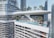 A modern skyscraper features a luxurious rooftop with an expansive infinity pool and lounging area. The building's curved glass and steel design is surrounded by tall city skyscrapers. Palm trees and sun loungers are scattered around the pool, where people are relaxing and enjoying the view.