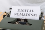 A vintage typewriter with a sheet of paper in the carriage displaying the phrase 'DIGITAL NOMADISM'. The typewriter has a dark green body and metal components, set against a light background.