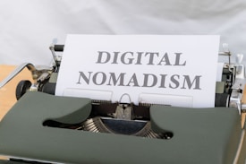 A vintage typewriter with a sheet of paper in the carriage displaying the phrase 'DIGITAL NOMADISM'. The typewriter has a dark green body and metal components, set against a light background.
