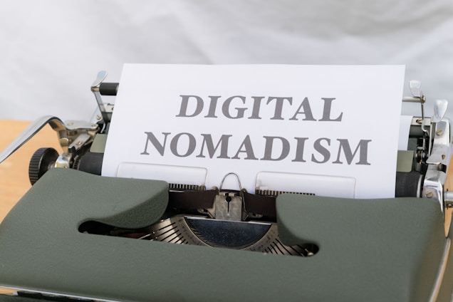 A vintage typewriter with a sheet of paper in the carriage displaying the phrase 'DIGITAL NOMADISM'. The typewriter has a dark green body and metal components, set against a light background.