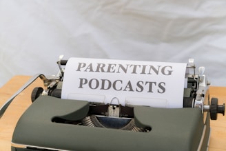 A close-up of an old-fashioned typewriter featuring a sheet of paper with the words 'PARENTING PODCASTS' typed on it. The typewriter is placed on a wooden surface and the background is a plain, neutral color.