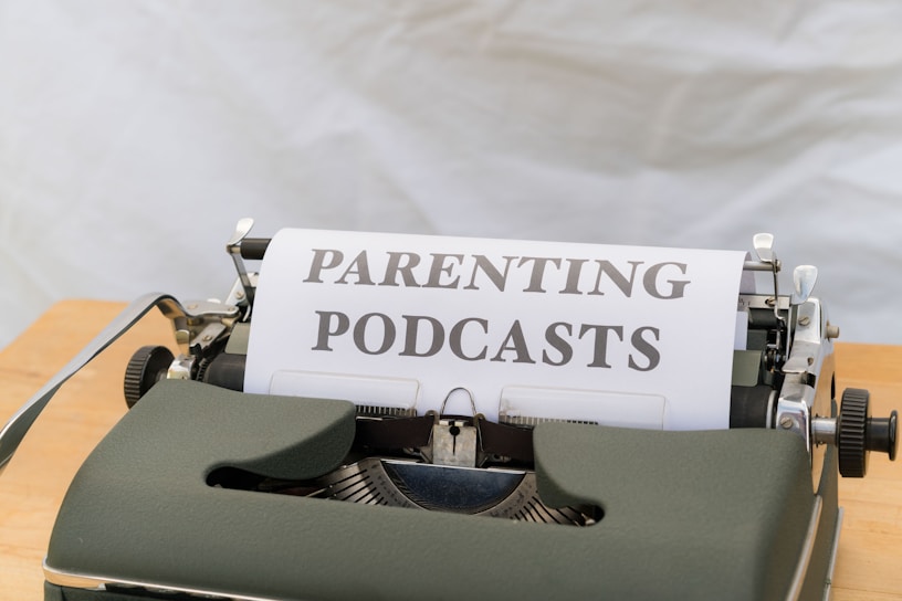 A close-up of an old-fashioned typewriter featuring a sheet of paper with the words 'PARENTING PODCASTS' typed on it. The typewriter is placed on a wooden surface and the background is a plain, neutral color.