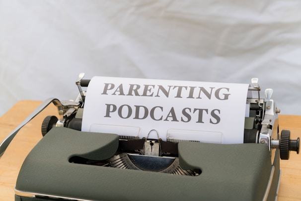 A close-up of an old-fashioned typewriter featuring a sheet of paper with the words 'PARENTING PODCASTS' typed on it. The typewriter is placed on a wooden surface and the background is a plain, neutral color.