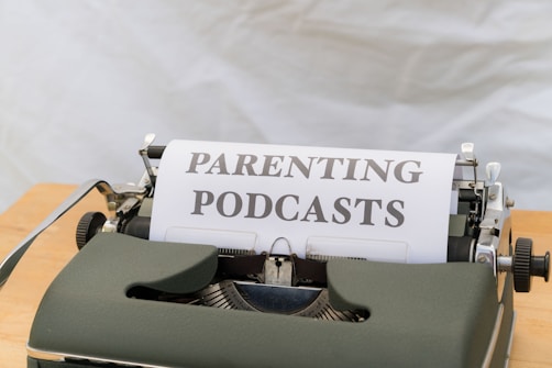 A close-up of an old-fashioned typewriter featuring a sheet of paper with the words 'PARENTING PODCASTS' typed on it. The typewriter is placed on a wooden surface and the background is a plain, neutral color.