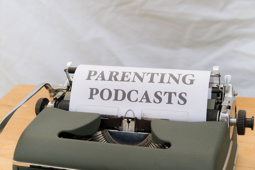A close-up of an old-fashioned typewriter featuring a sheet of paper with the words 'PARENTING PODCASTS' typed on it. The typewriter is placed on a wooden surface and the background is a plain, neutral color.