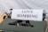 A close-up view of a vintage typewriter with a sheet of paper displaying the text 'LOVE BOMBING' in bold, capital letters. The typewriter is dark green and black, positioned against a neutral, light-colored background.
