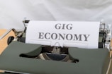 A vintage typewriter with a sheet of paper displaying the words 'GIG ECONOMY' in bold uppercase letters. The typewriter is green and sits on a light wooden surface.