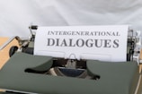 A vintage typewriter with a piece of paper inserted displays the title 'Intergenerational Dialogues' in bold text. The machine features a classic design with a green cover and visible keys, set against a blurred background.