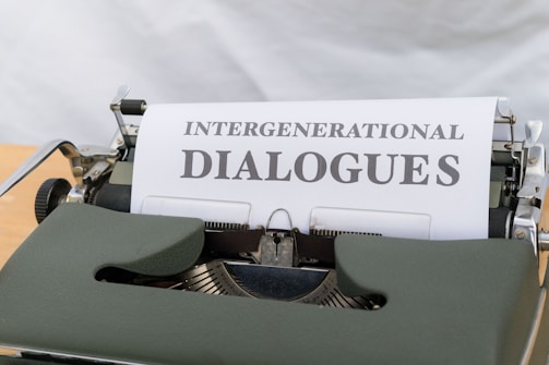A vintage typewriter with a piece of paper inserted displays the title 'Intergenerational Dialogues' in bold text. The machine features a classic design with a green cover and visible keys, set against a blurred background.