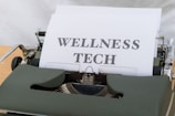 A vintage typewriter holds a sheet of paper with the words 'WELLNESS TECH' typed in large, bold letters. The typewriter is in a shade of olive green, with a focus on the metal and mechanical components. The paper is placed neatly in the roller, ready for typing more text.