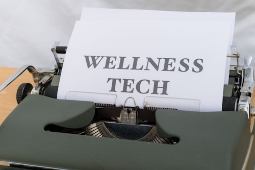 A vintage typewriter holds a sheet of paper with the words 'WELLNESS TECH' typed in large, bold letters. The typewriter is in a shade of olive green, with a focus on the metal and mechanical components. The paper is placed neatly in the roller, ready for typing more text.